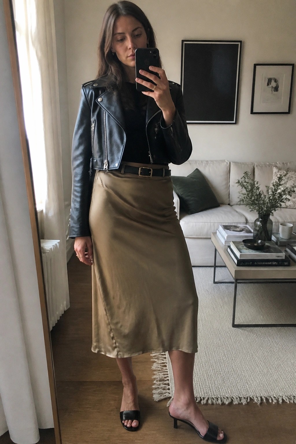Woman in black leather jacket and beige satin skirt taking mirror selfie in living room.