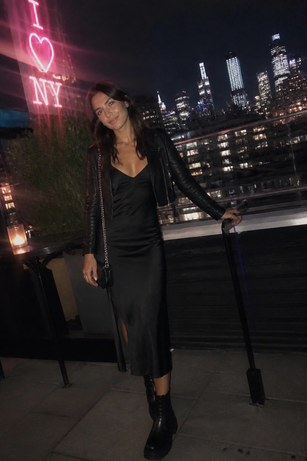Woman in black satin dress and leather jacket on NYC rooftop at night with I Love NY sign