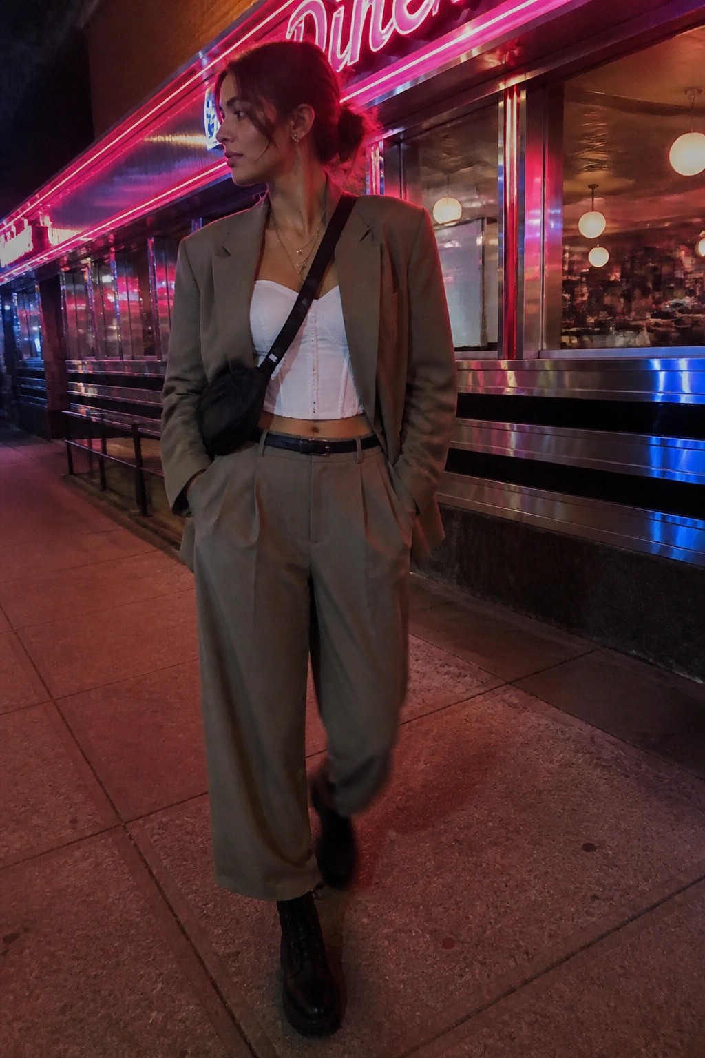 Woman in sage green suit and white corset outside neon-lit diner at night.