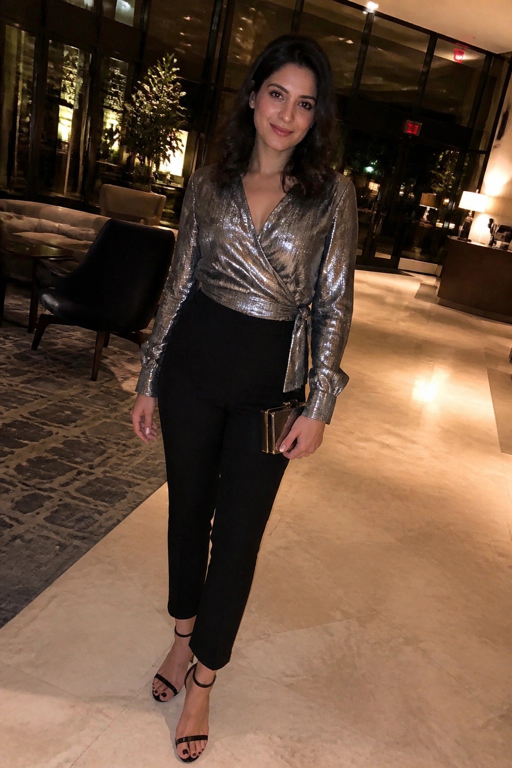 Woman in silver sequined wrap top and black pants holding gold clutch in lobby
