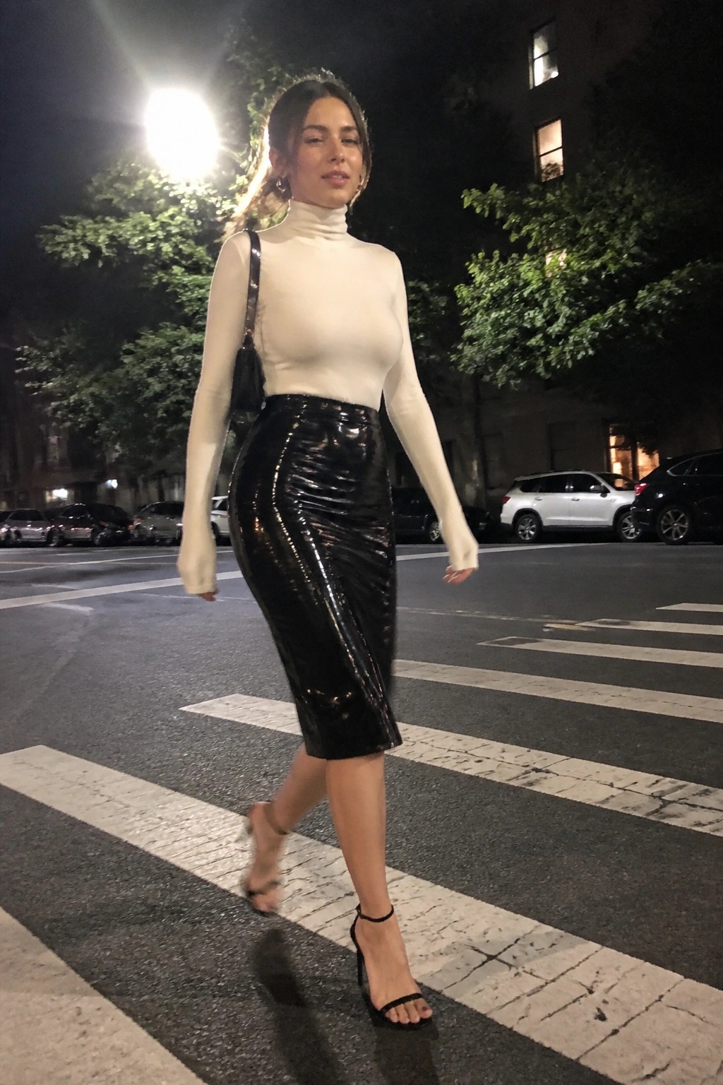 Woman crossing street at night in fitted white turtleneck top with sheer sleeves, glossy black patent midi skirt, black strappy high heels and small black crossbody bag.