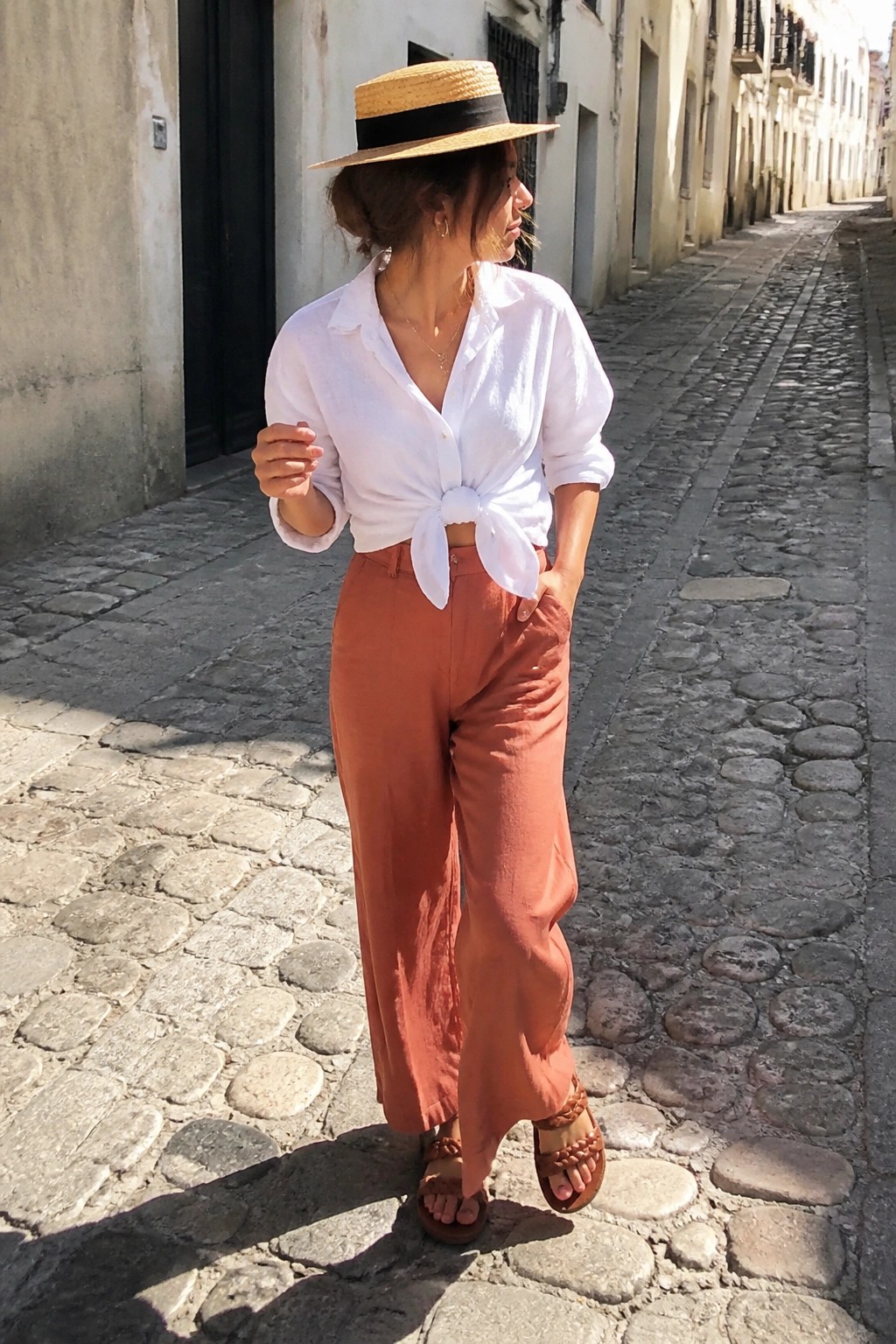 Woman wearing a white button-down shirt tied in a knot at the waist with rust-colored wide-leg pants, a straw hat with black band, and brown woven sandals standing on a cobblestone street.