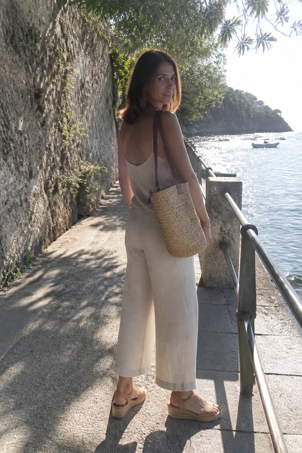 Woman in beige jumpsuit with straw bag on sunny coastal path by sea