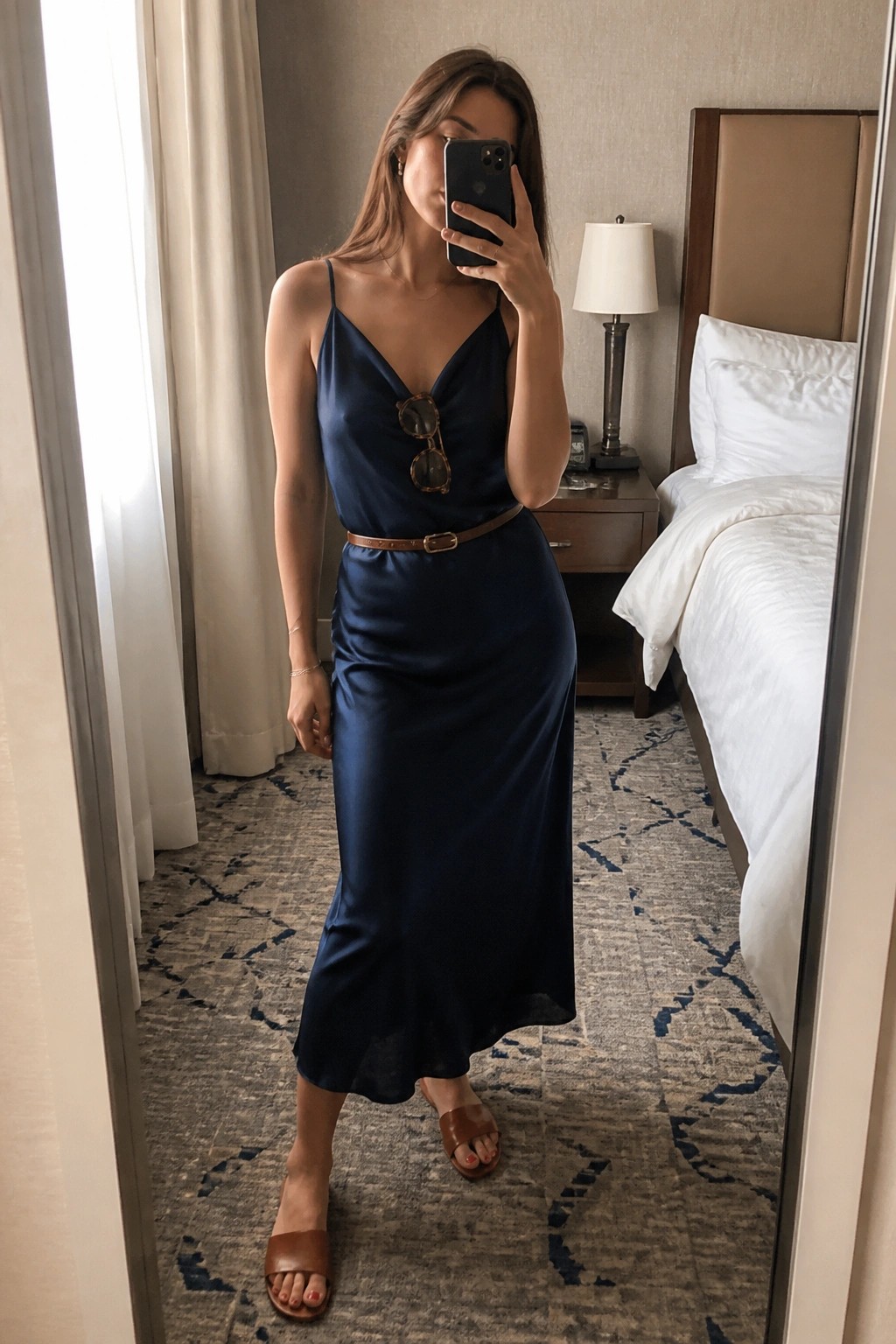 Woman in navy satin dress with brown belt taking mirror selfie in hotel room.
