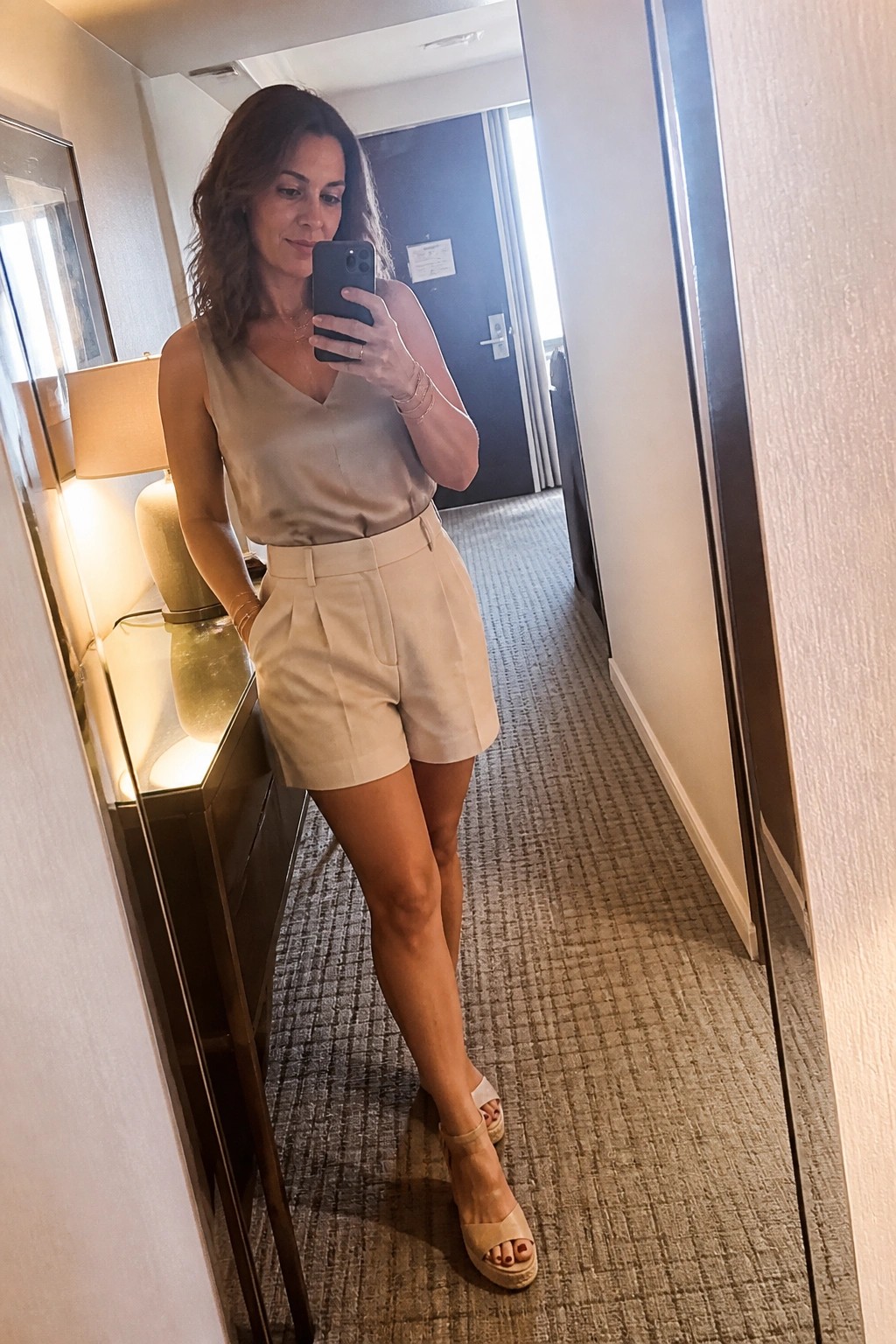 Woman in beige satin top and shorts taking mirror selfie in hotel hallway.