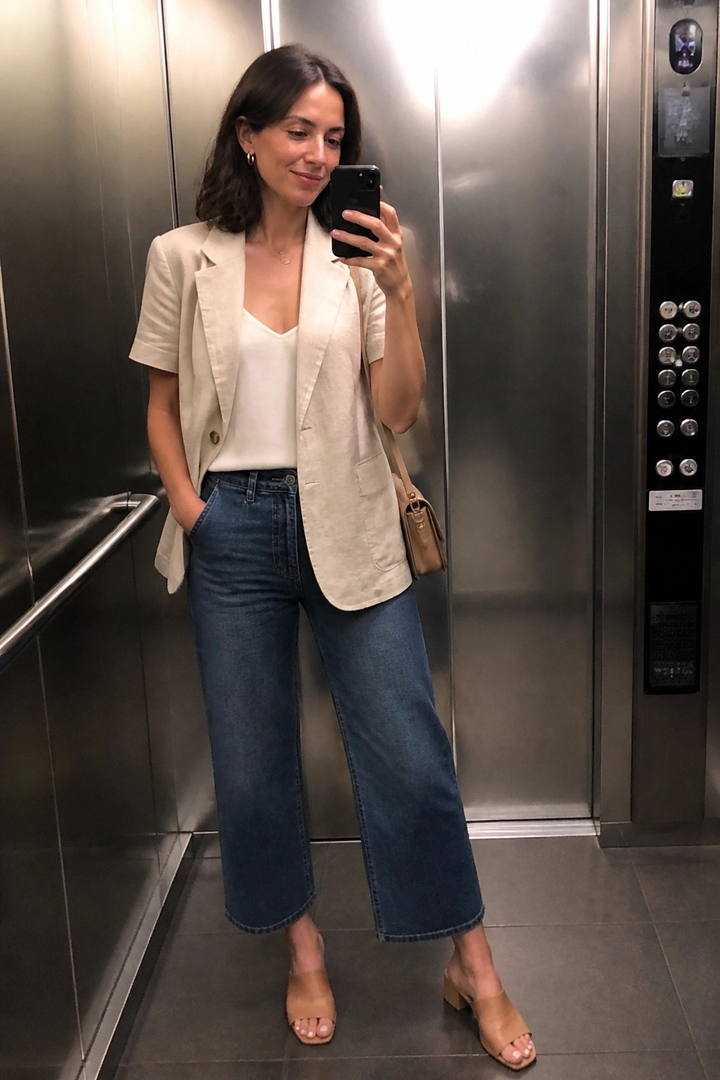 Woman in a cream short-sleeve blazer over a white camisole paired with blue wide-leg cropped jeans and tan block-heel mules, holding a phone for a mirror selfie inside an elevator.