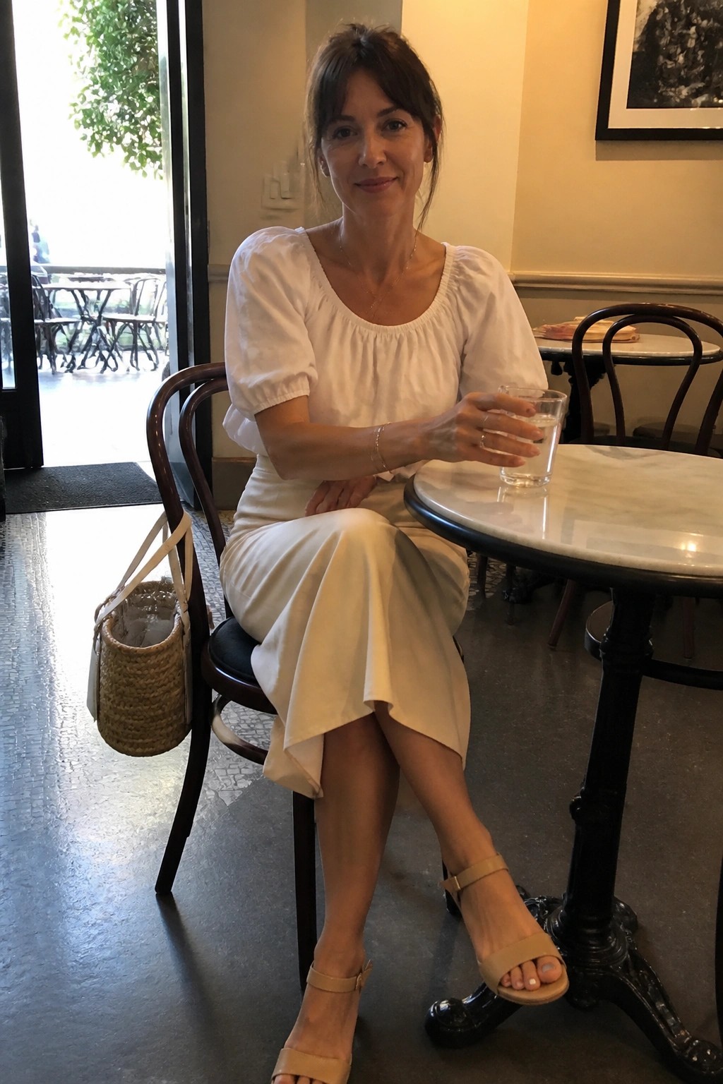 Woman in white top and skirt at marble cafe table holding water glass
