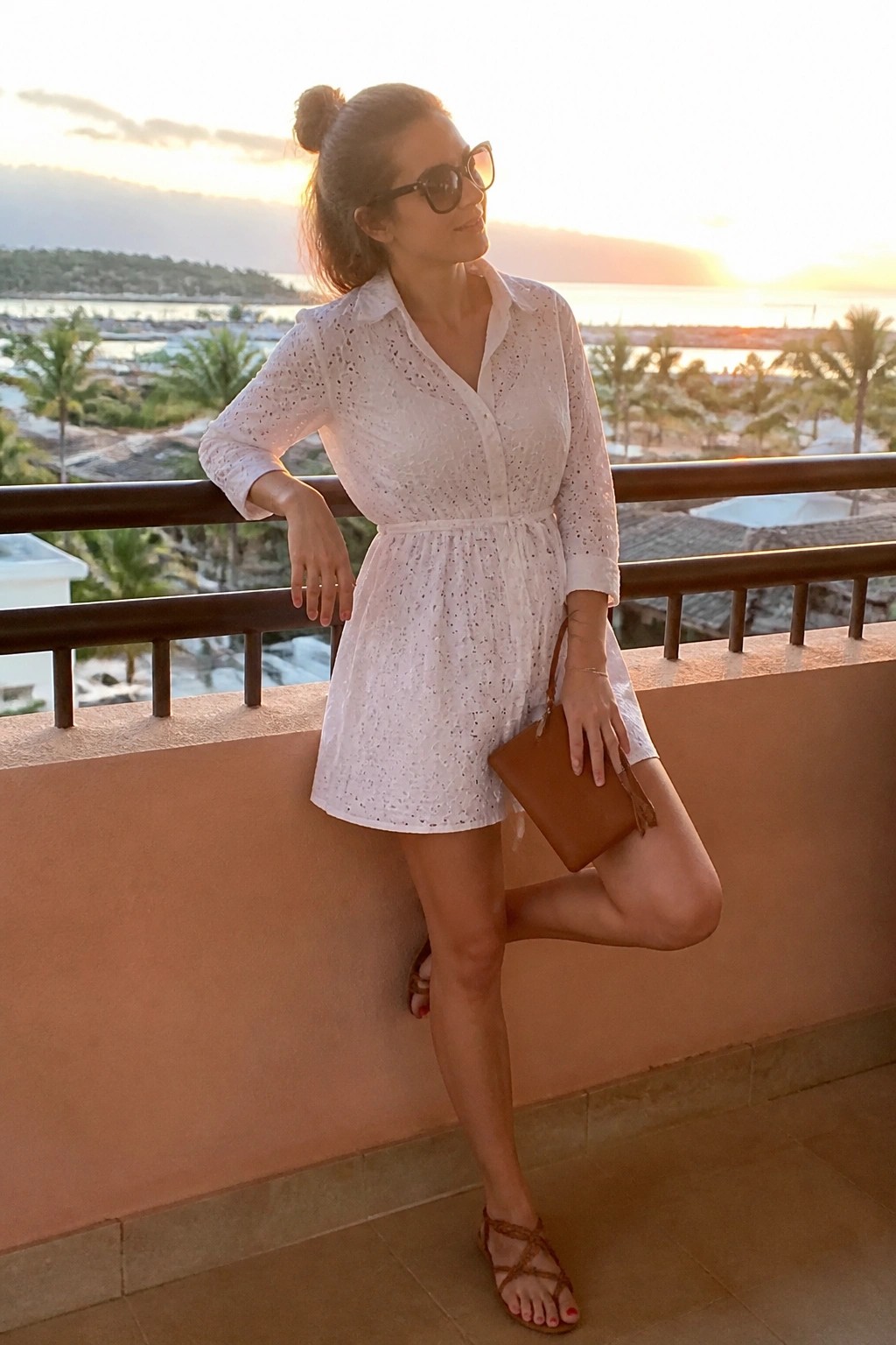 Woman in white lace dress posing on balcony at golden hour sunset.