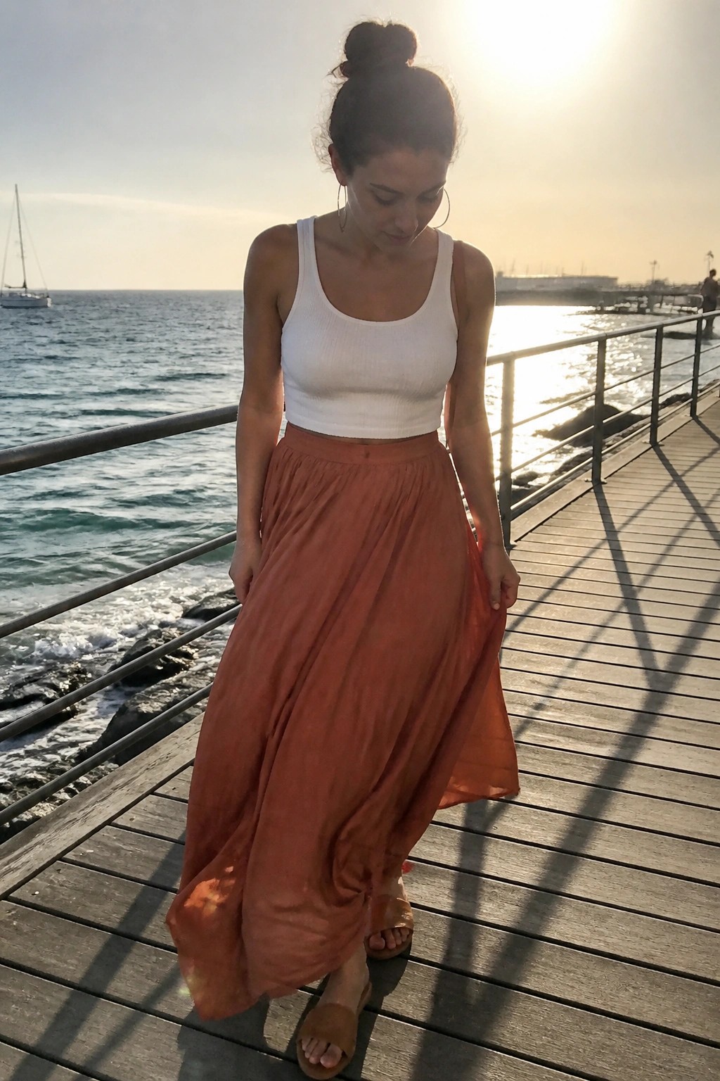Woman in white crop top and flowing orange skirt on wooden pier by sea at sunset
