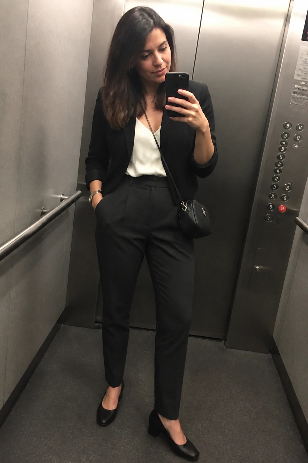 Woman standing in an elevator wearing a black blazer over a white camisole with matching black trousers, black block heel shoes, and a small black crossbody bag while holding a phone.