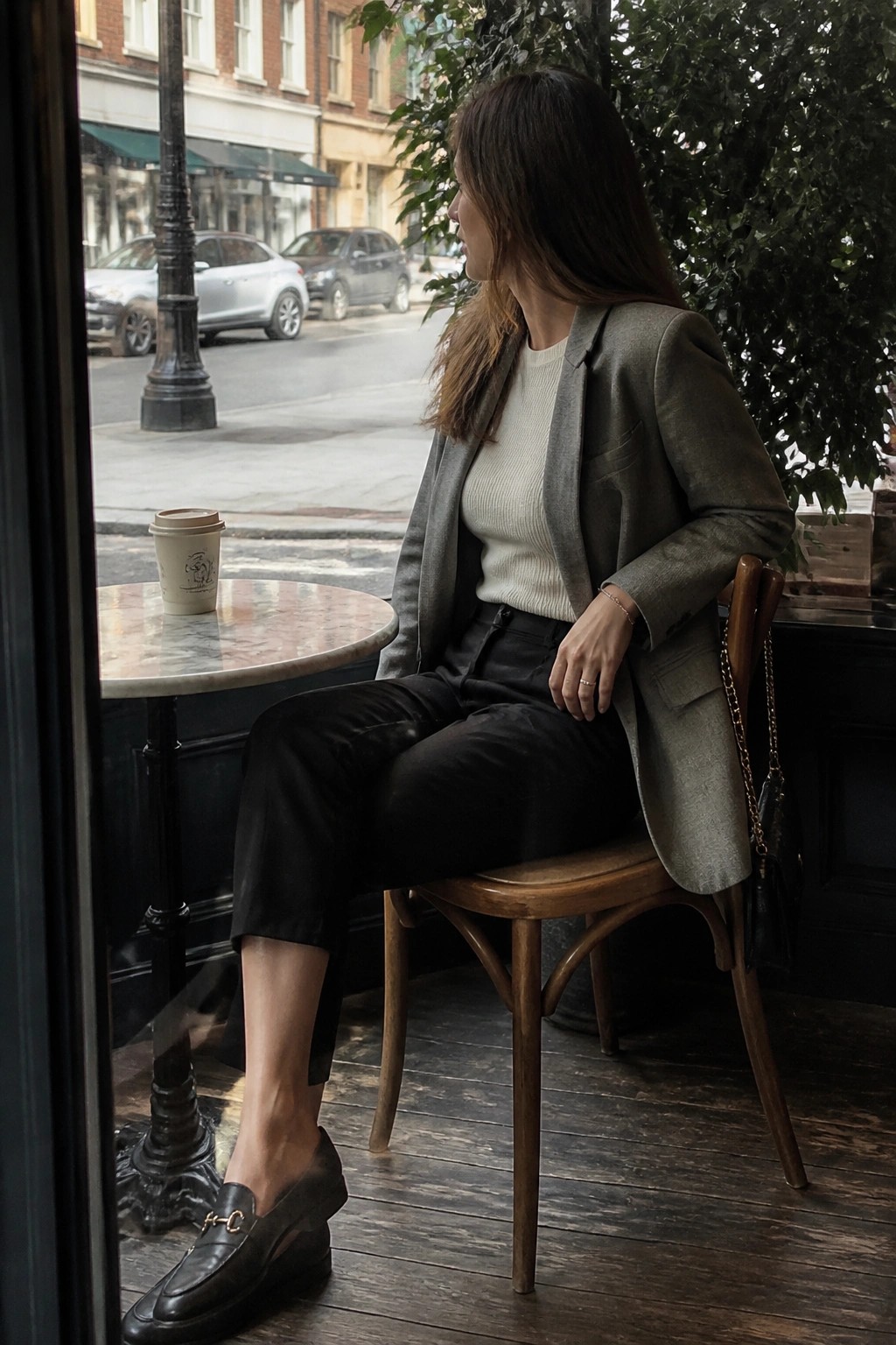 Woman in gray blazer and black pants seated at cafe window with coffee cup