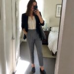 Woman in navy blazer and gray trousers taking mirror selfie with black handbag.