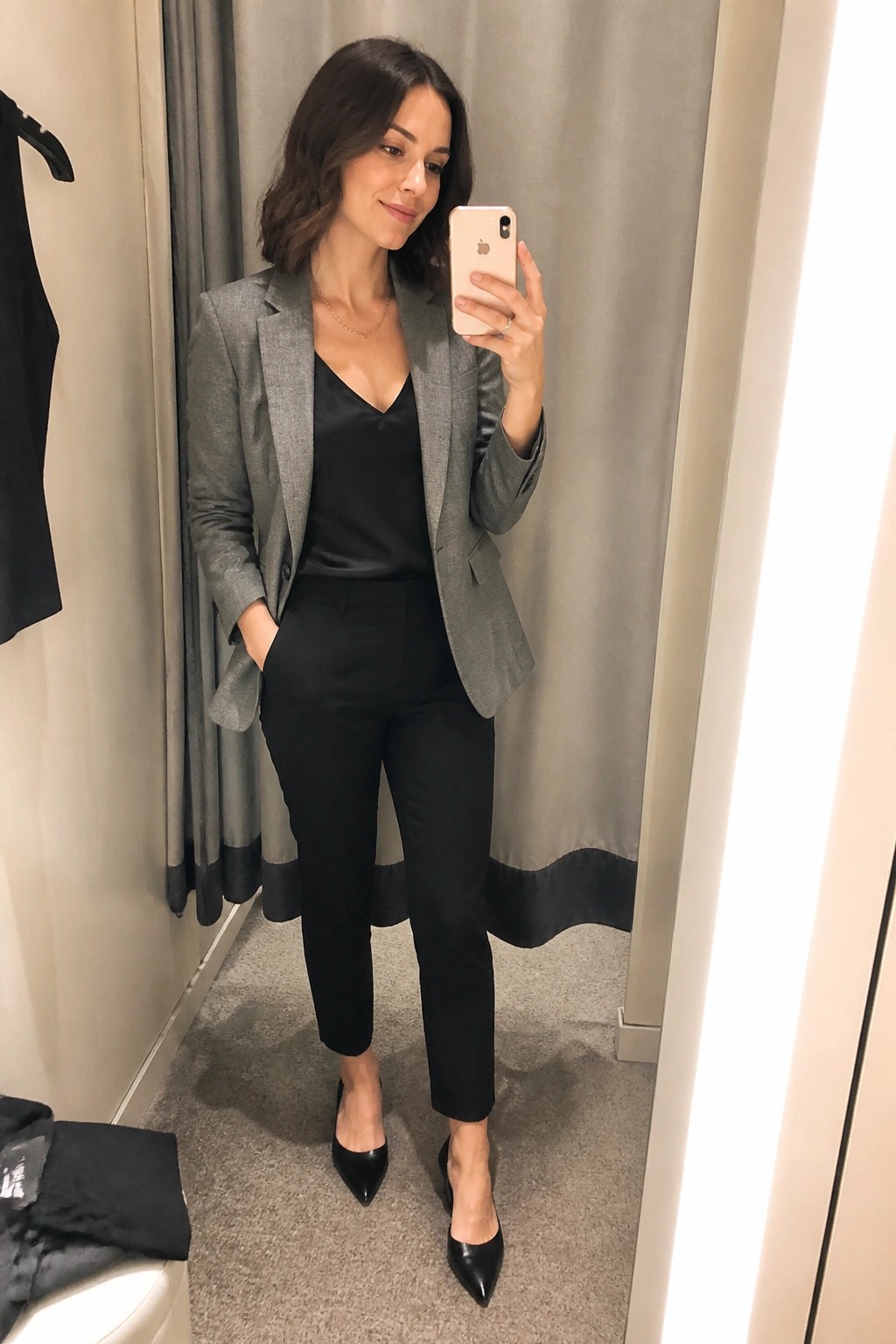 Woman wearing a grey tailored blazer over a black camisole with matching black trousers and pointed black heels in a mirror selfie.