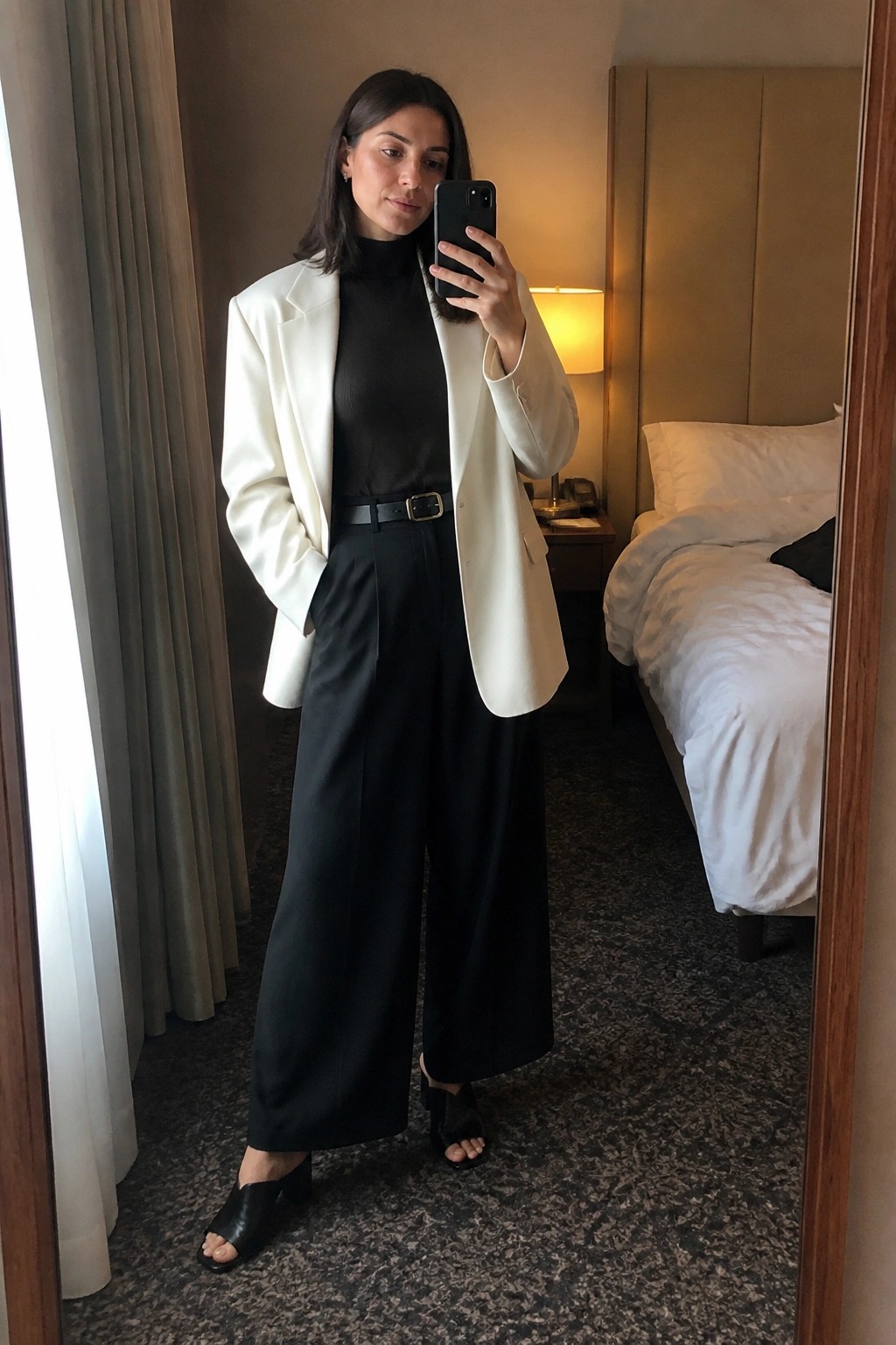 Woman in oversized white blazer over black turtleneck with black wide leg trousers belted at waist and black heeled sandals standing in hotel room mirror selfie.