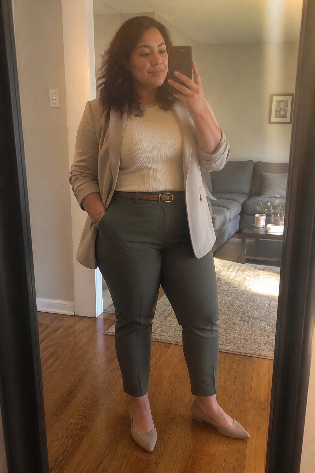 Woman in plus size work outfit with beige blazer, cream ribbed sweater, green tailored pants, brown belt, and beige pointed flats taking a mirror selfie.
