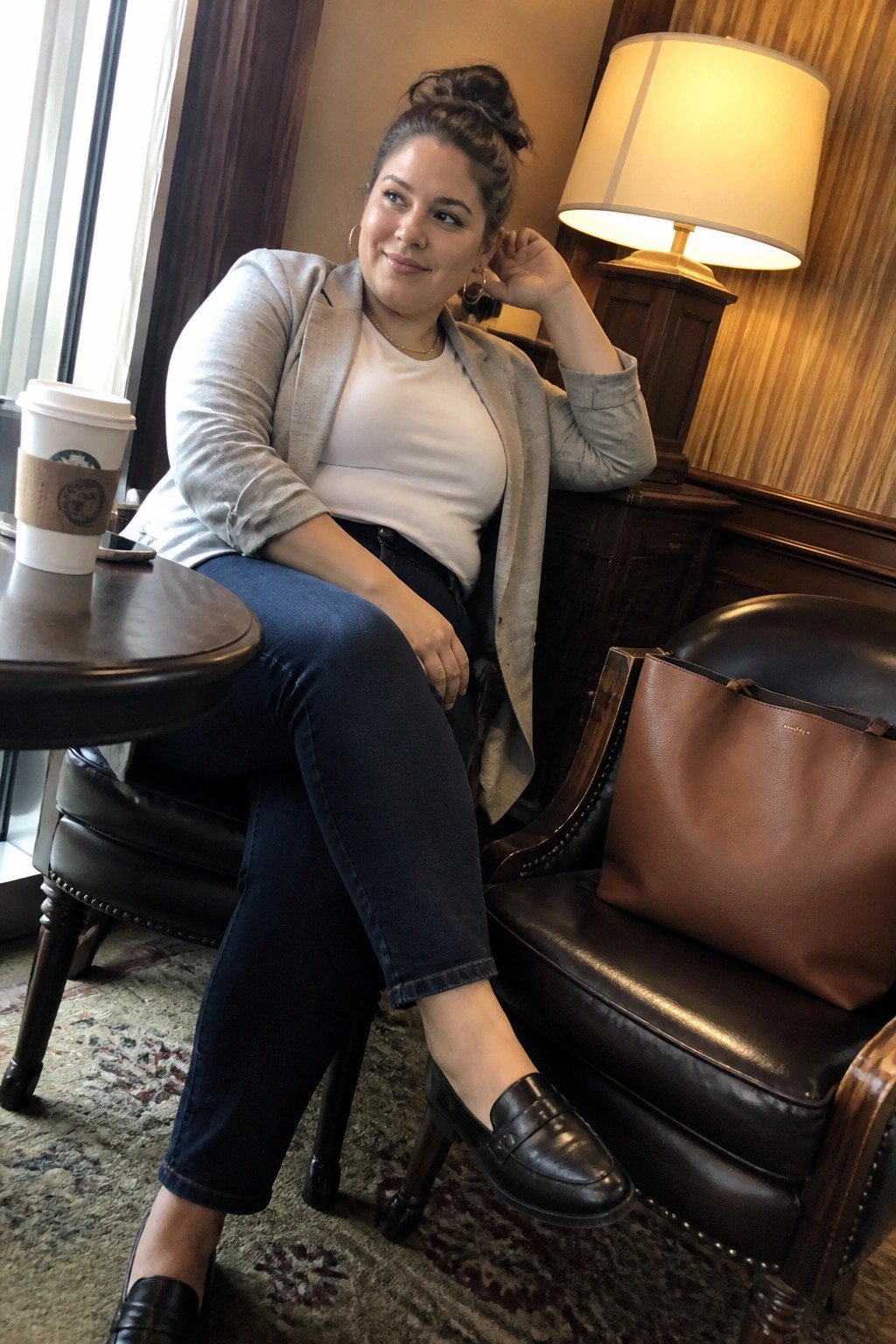 Woman in gray blazer and jeans seated by brown leather bag and Starbucks cup.