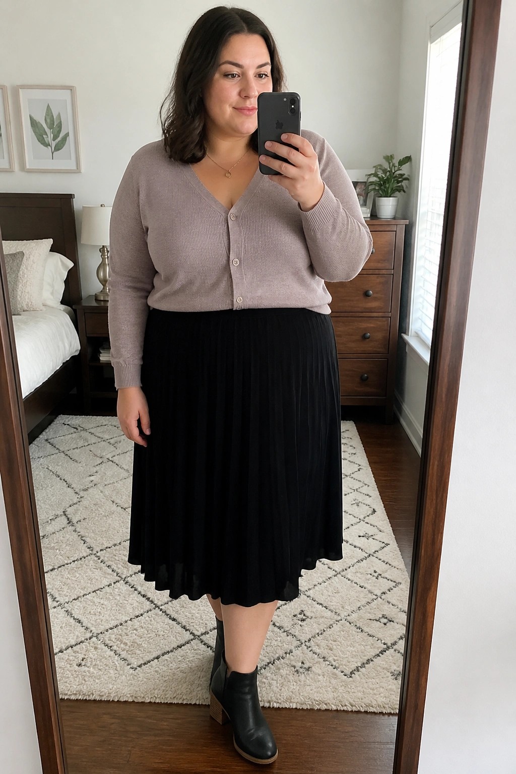 Plus size woman wearing a light purple knit cardigan buttoned over a black pleated midi skirt with black ankle boots, holding a phone for a mirror selfie in a bedroom setting.