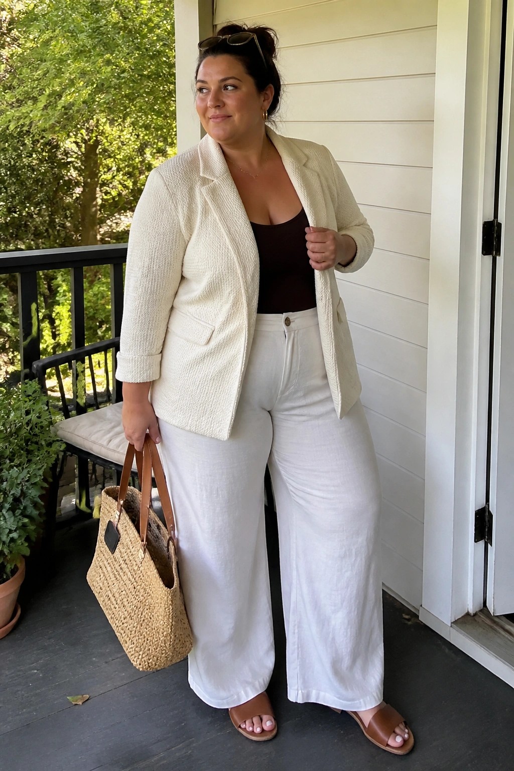 Woman in cream blazer and white wide-leg pants holding woven tote on porch