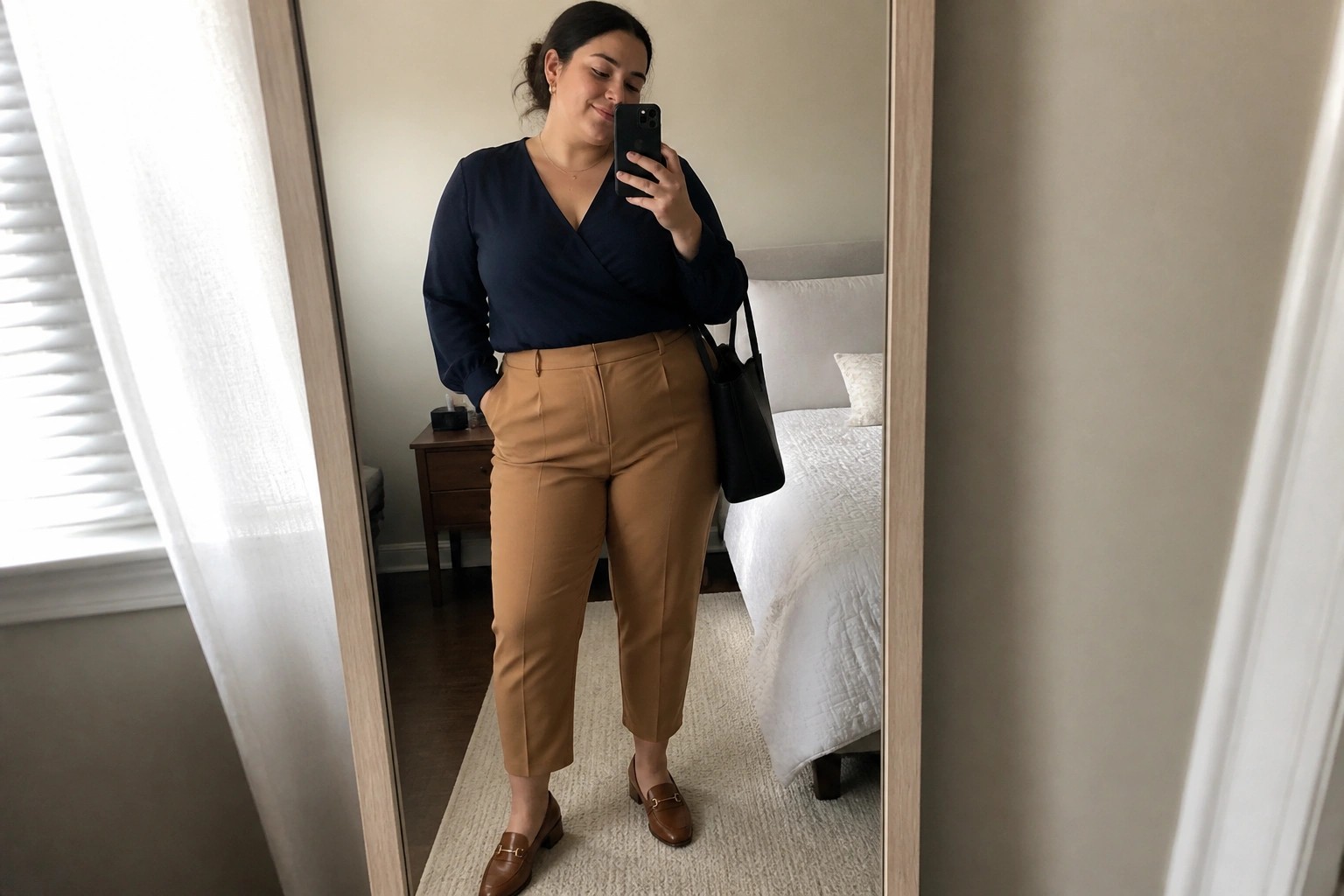 Woman in navy wrap top and tan pants taking bedroom mirror selfie