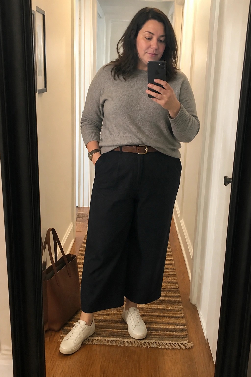 Woman in gray sweater and black pants taking mirror selfie in hallway.