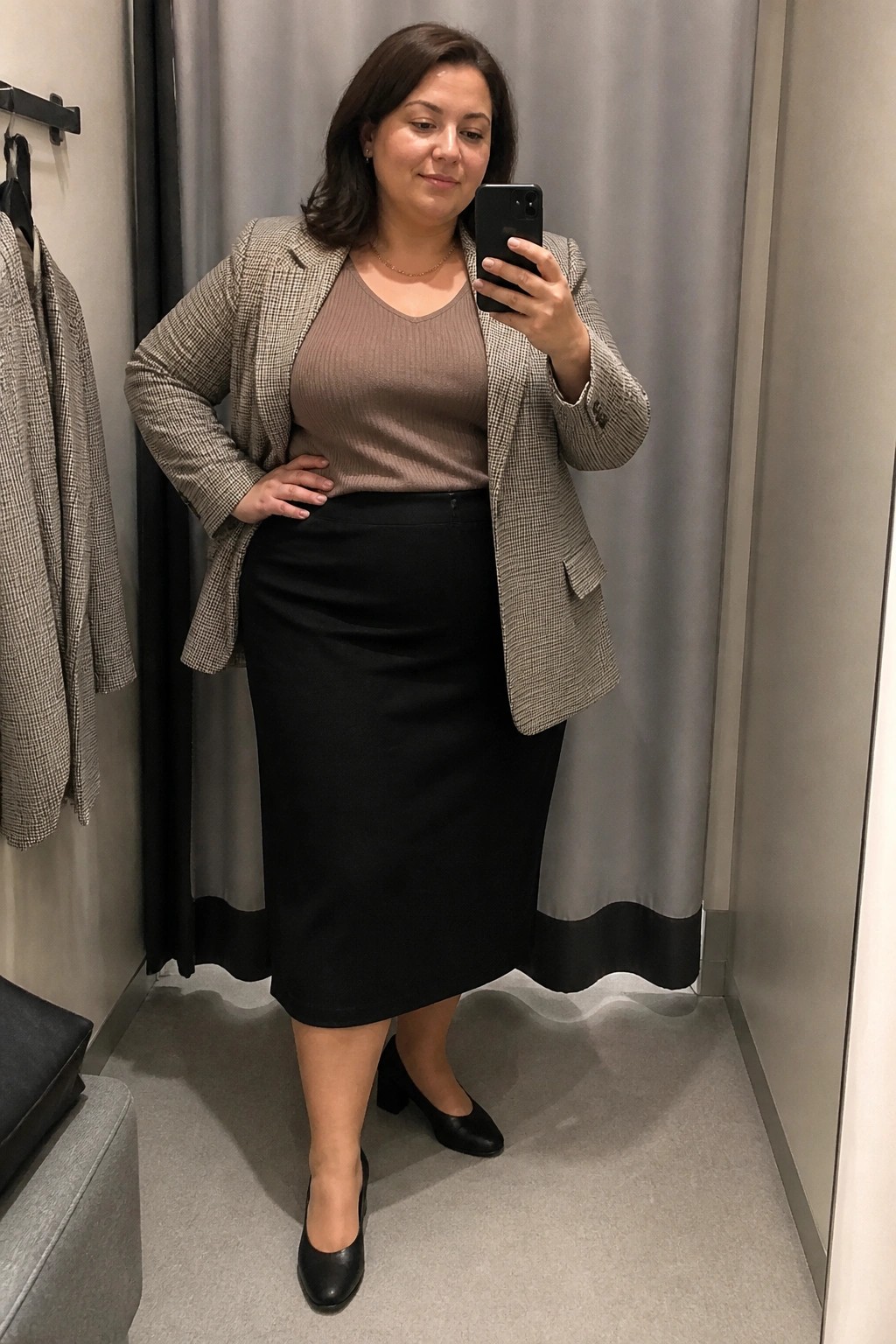 Woman in checkered blazer and black skirt taking mirror selfie in fitting room.