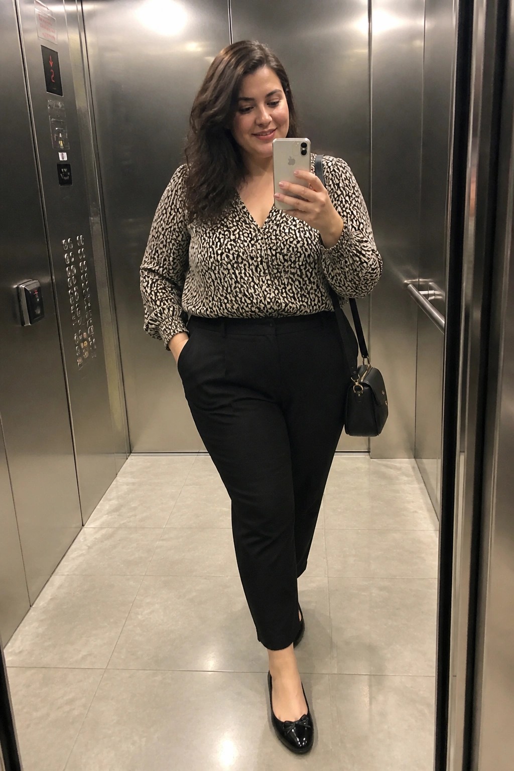 Plus size woman wearing black and white patterned long sleeve blouse with black trousers and black flats while holding a phone in an elevator mirror selfie.