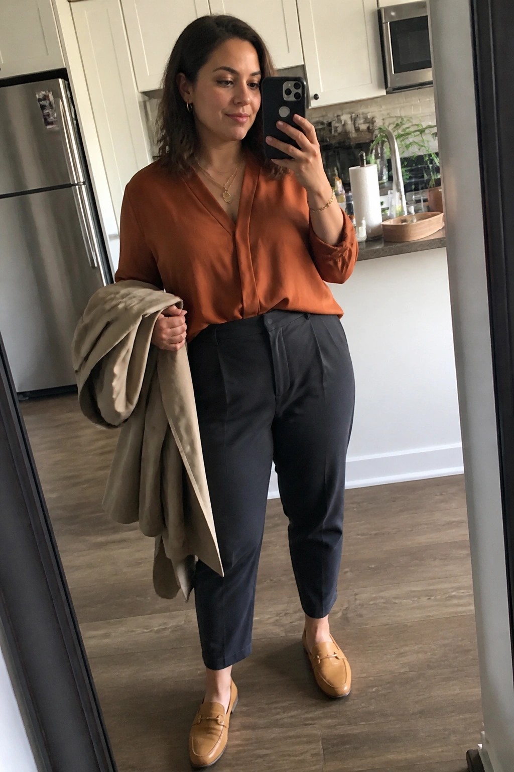 Woman in orange blouse and gray pants holding beige jacket taking kitchen mirror selfie