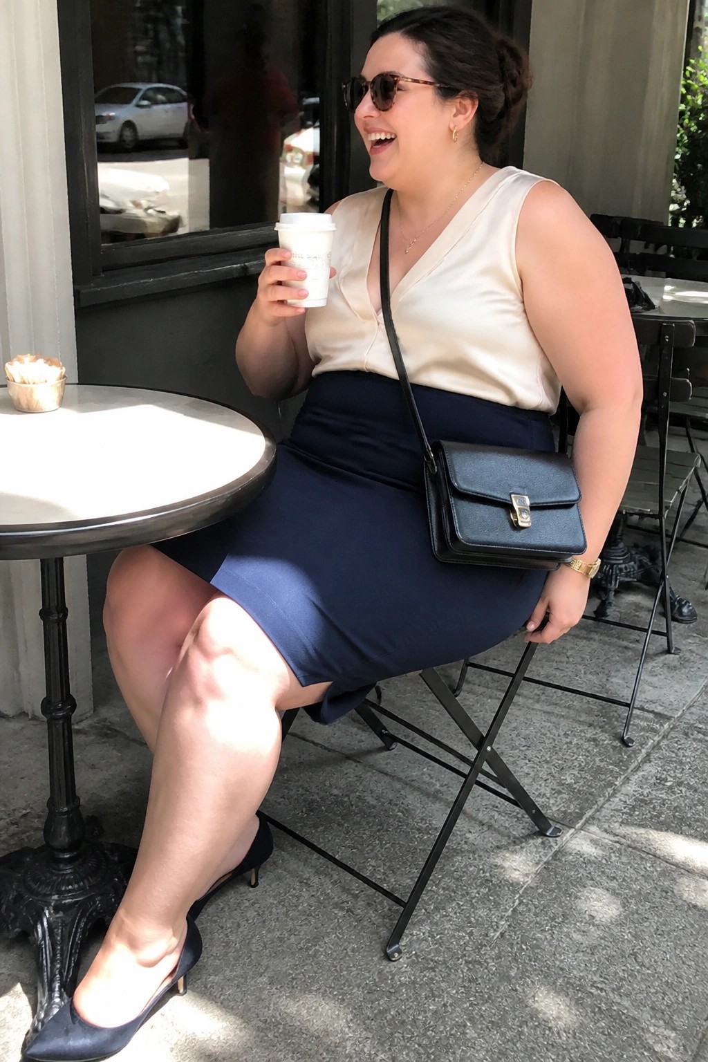 Woman in cream top and navy skirt holding coffee at outdoor cafe.