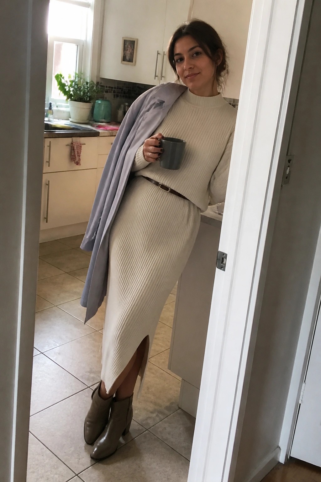Woman in cream ribbed dress with lavender coat over shoulders holding gray mug in kitchen