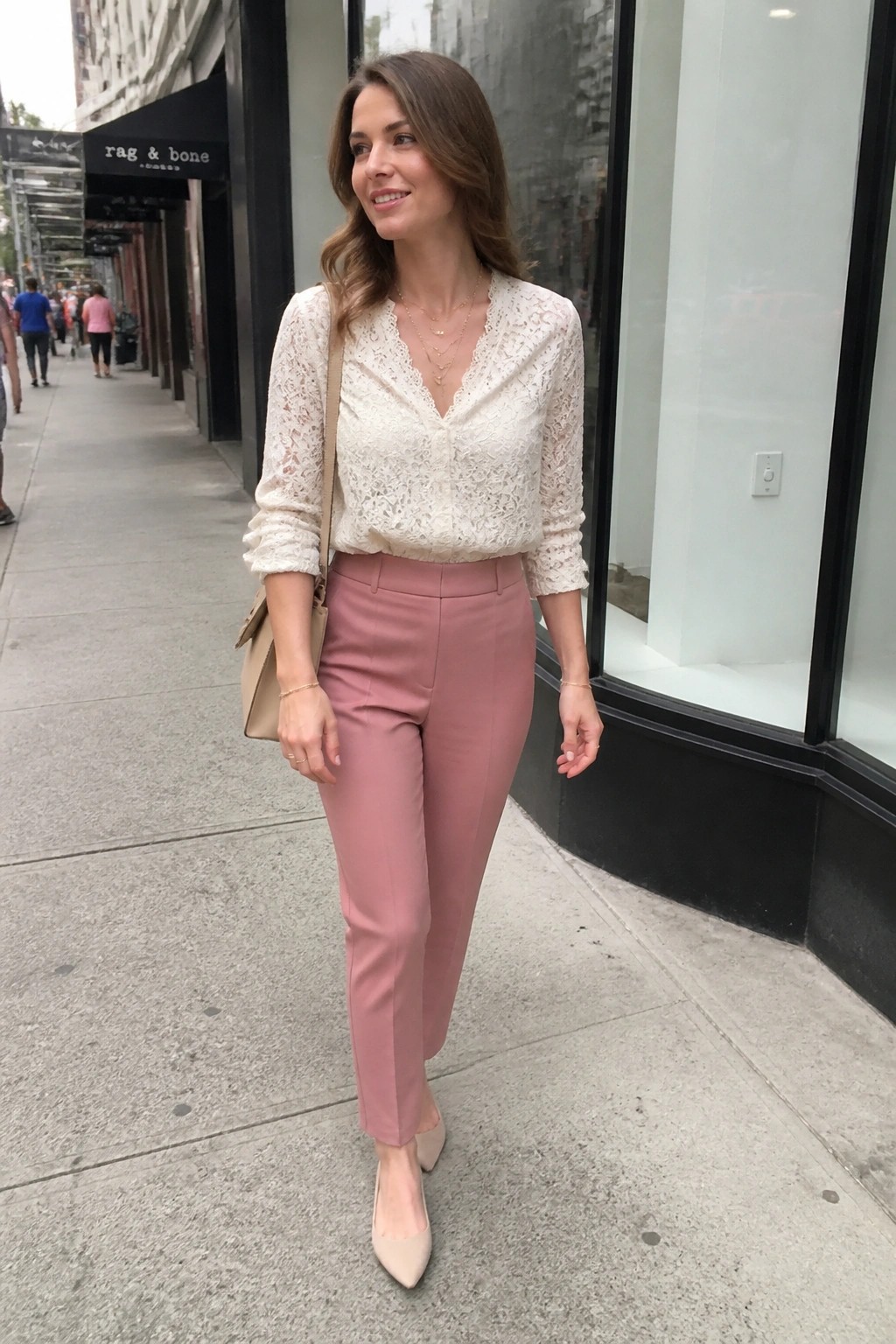 Woman in white lace blouse and pink trousers on urban sidewalk with beige bag.