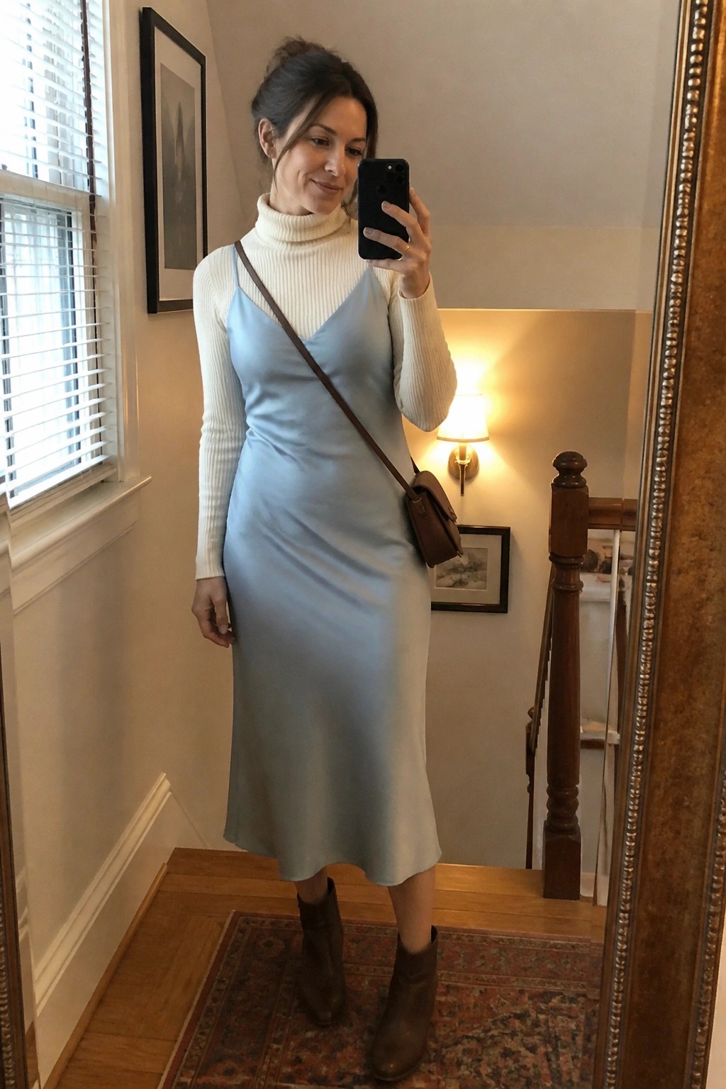 Woman in light blue slip dress over white turtleneck taking mirror selfie on staircase