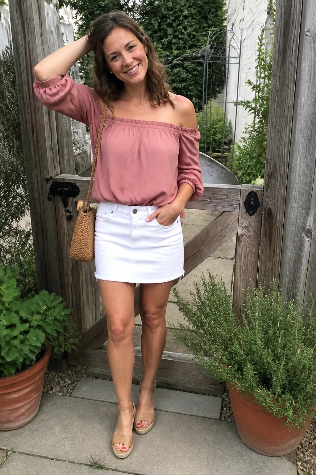 Woman in pink off-shoulder top and white mini skirt by wooden gate with plants