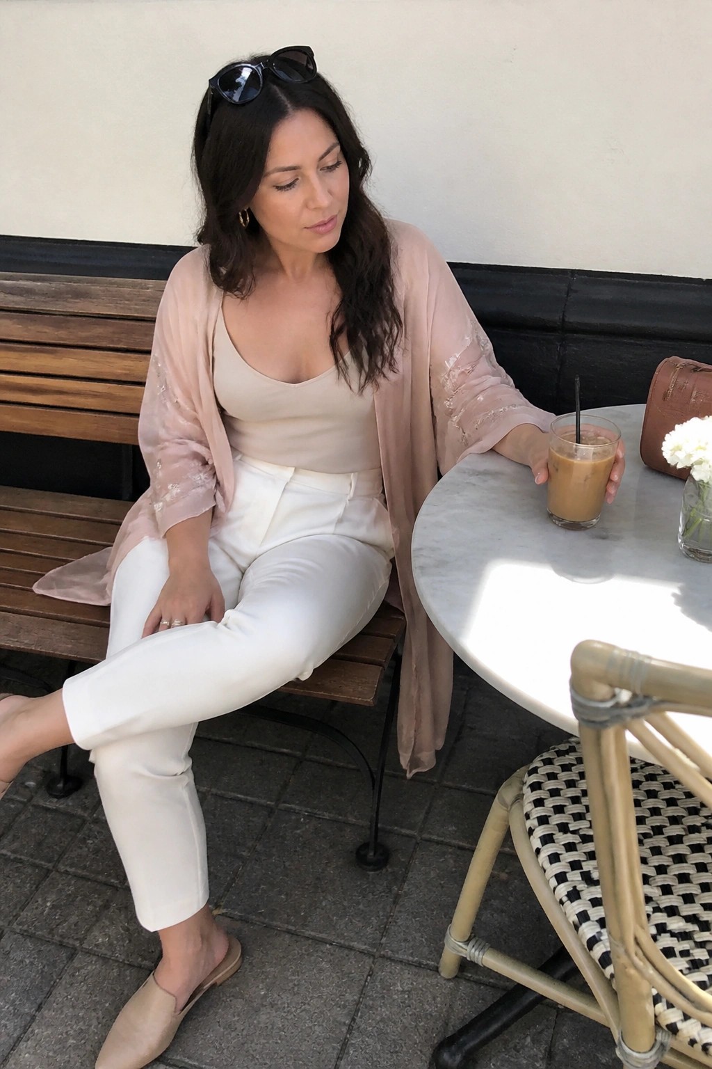 Woman wearing white high-waisted pants, cream camisole, and sheer blush pink duster with embroidered sleeves, paired with beige mules and sunglasses on her head while seated at an outdoor table.