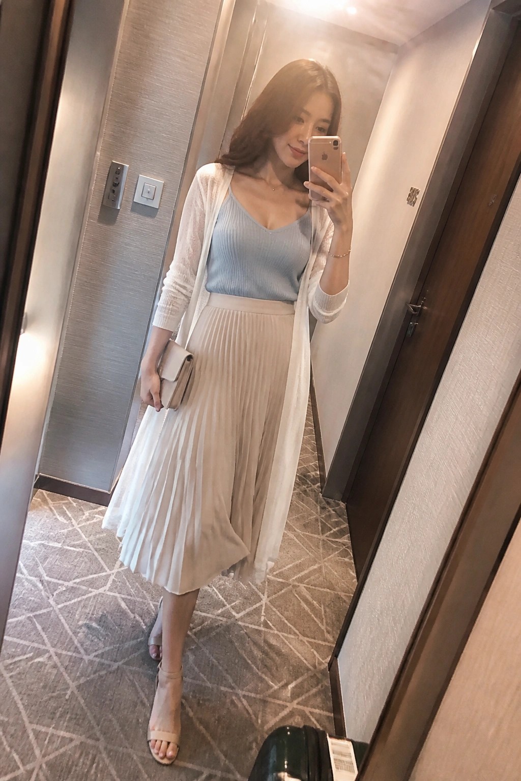 Woman taking a mirror selfie in a hallway wearing a light blue ribbed camisole under a sheer white long cardigan paired with a beige pleated midi skirt and matching heeled sandals while holding a beige clutch and phone.