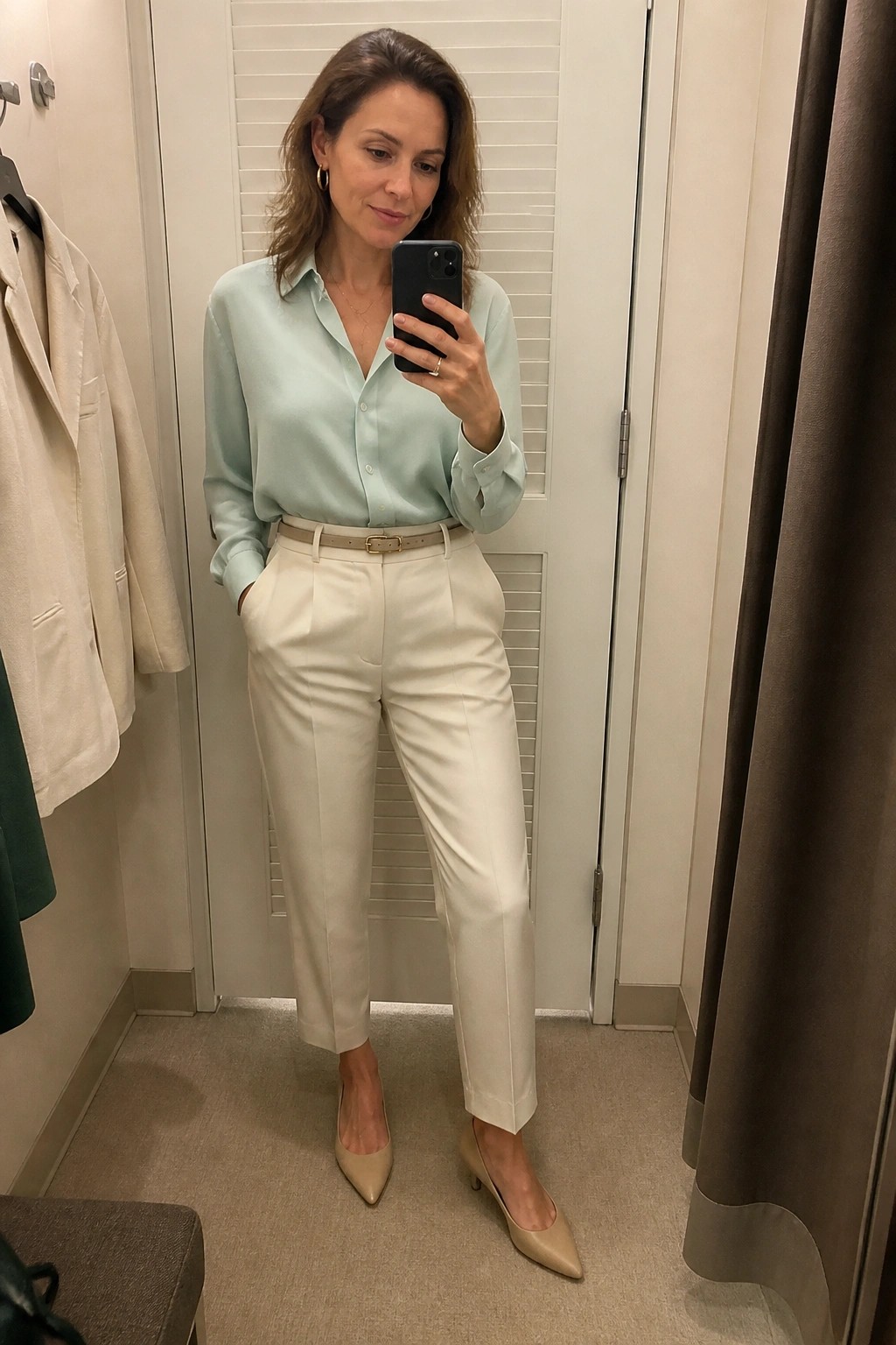 Woman wearing a mint green satin button up blouse tucked into high waisted cream trousers with a beige belt and matching pointed kitten heels while taking a mirror selfie in a fitting room.