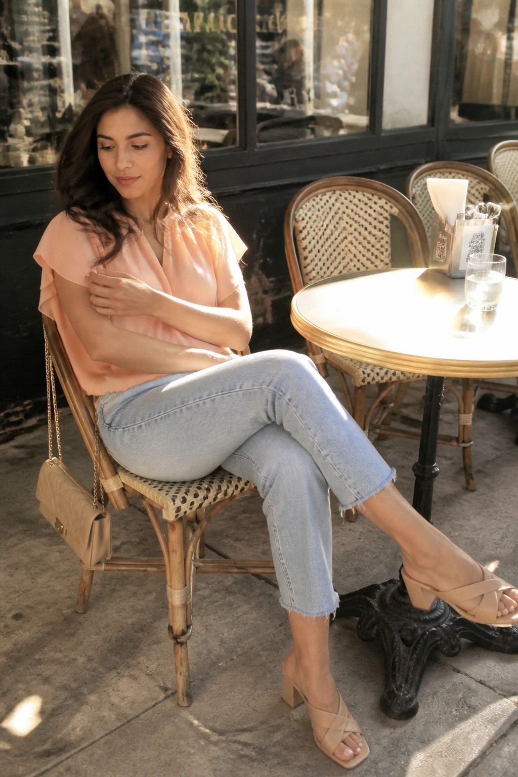 Woman in peach blouse and jeans seated at sunny outdoor cafe table with purse.