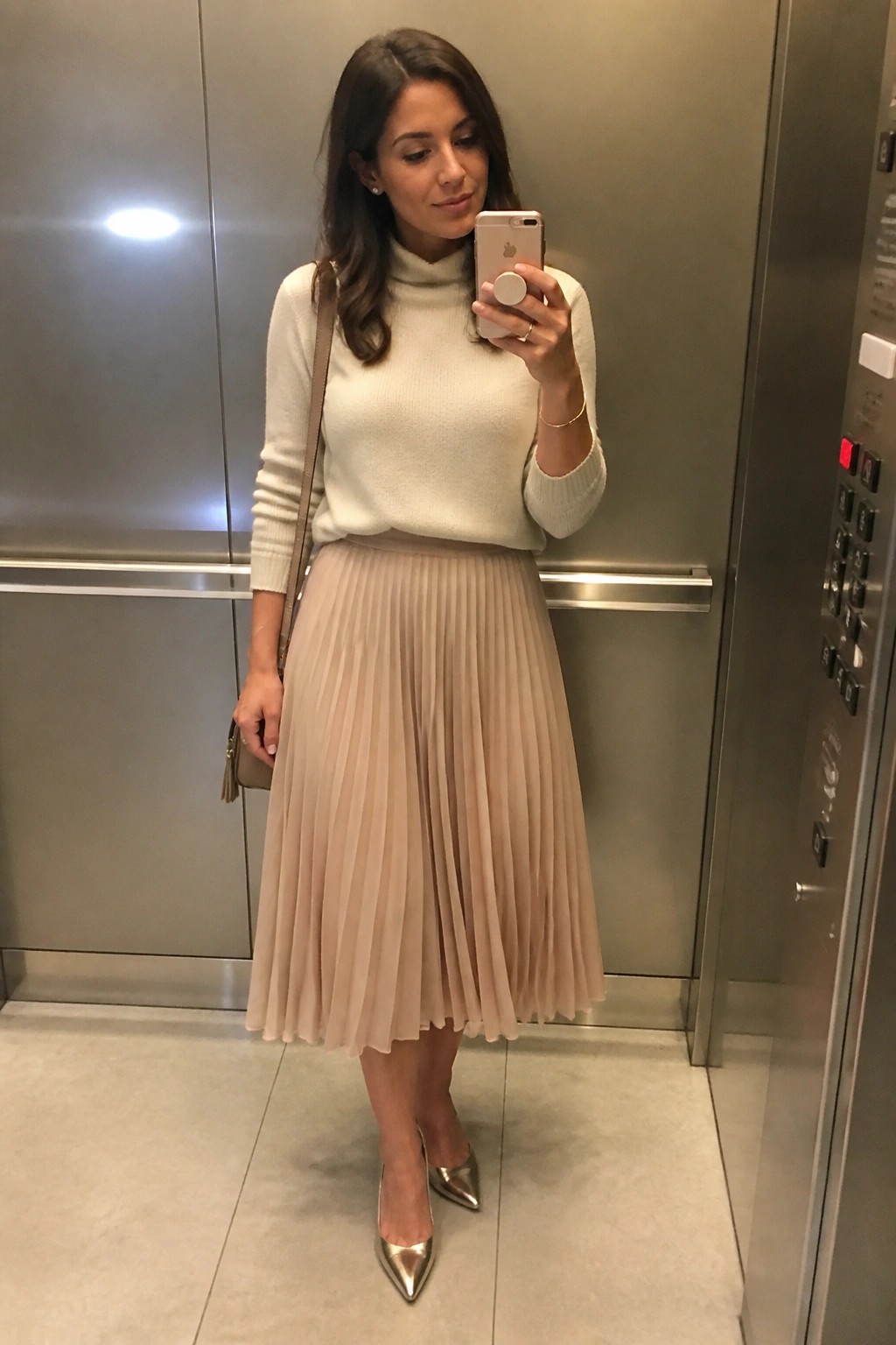 Woman in cream turtleneck and beige pleated skirt taking selfie in elevator