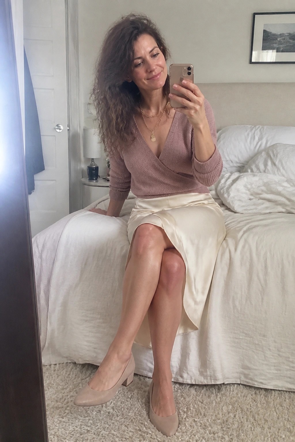 Woman in dusty rose wrap sweater and satin skirt taking bedroom selfie