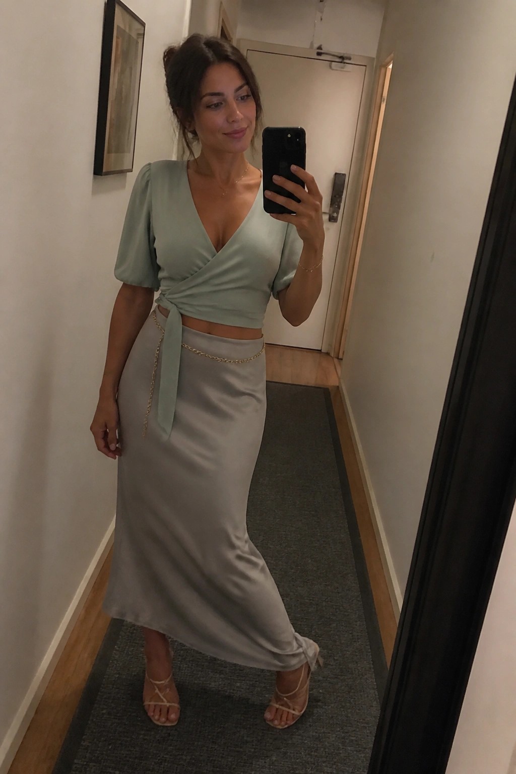 Woman in sage green satin wrap top and skirt taking mirror selfie in hallway.