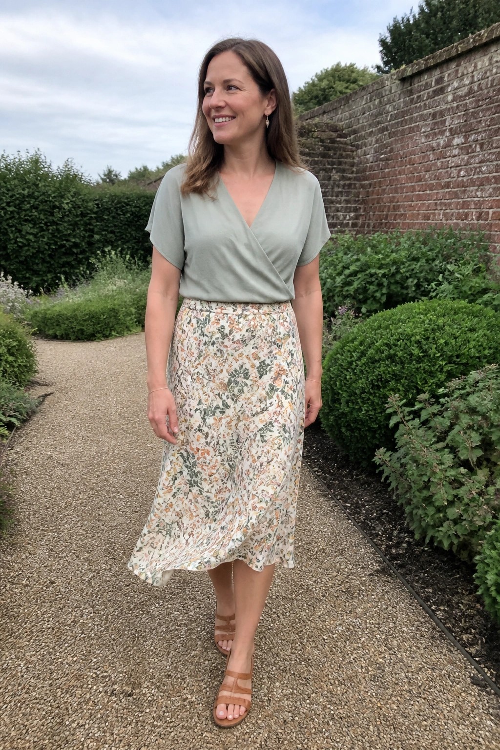 Woman wearing a sage green short-sleeve wrap top tucked into a cream floral midi skirt with orange and green accents, paired with tan strappy sandals and delicate gold jewelry.