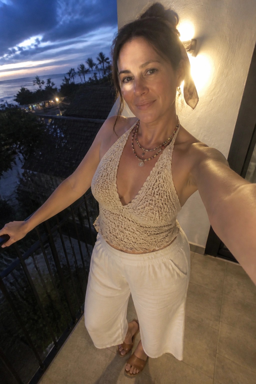Woman in white crochet halter top and pants taking balcony selfie at dusk overlooking ocean
