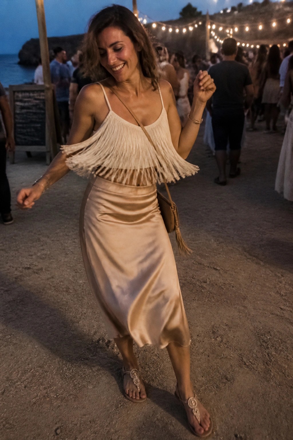 Woman in cream fringed crop top and matching satin midi skirt with jeweled thong sandals and small crossbody bag at a beach party.