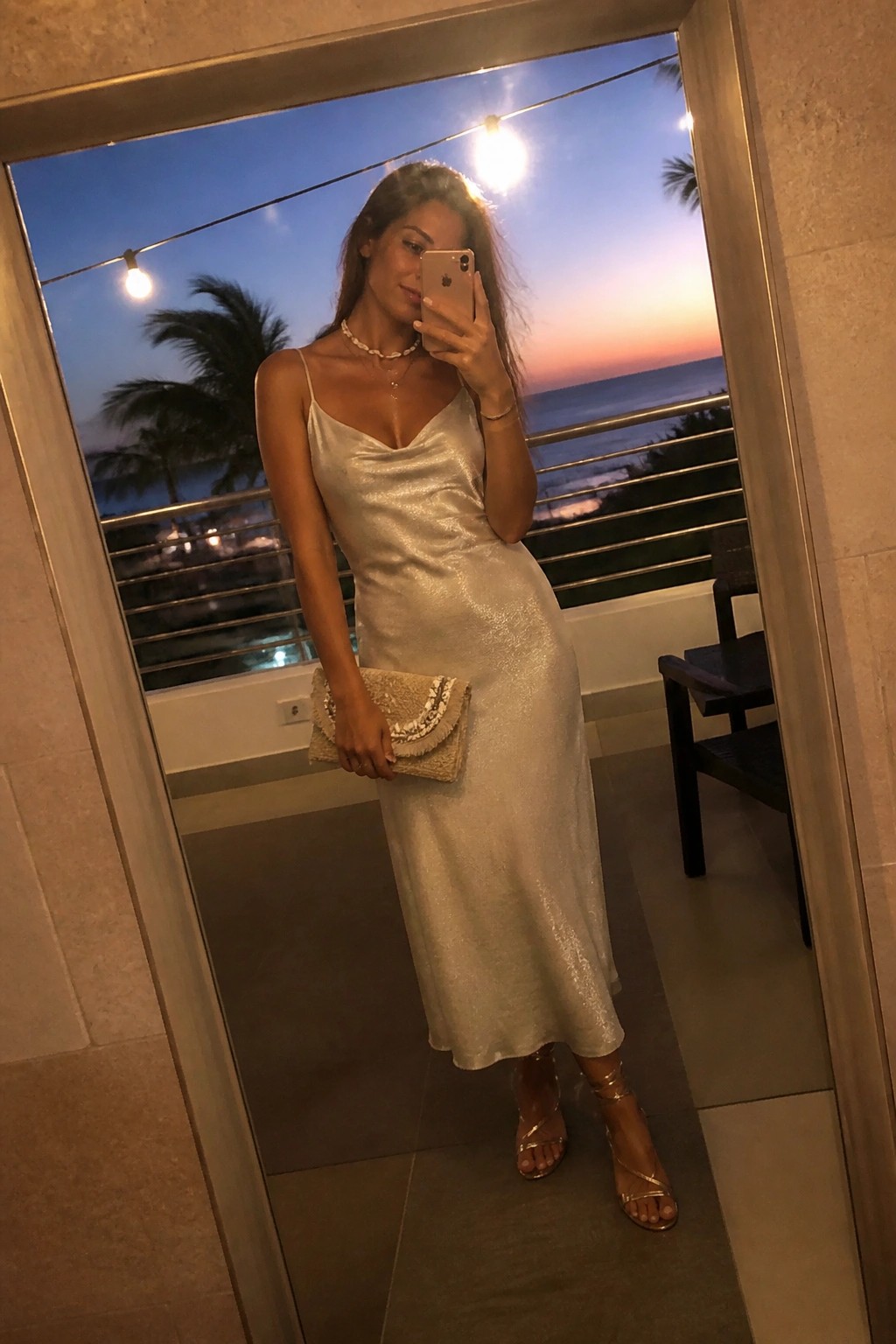 Woman in shimmering silver slip dress taking mirror selfie on ocean-view balcony at dusk