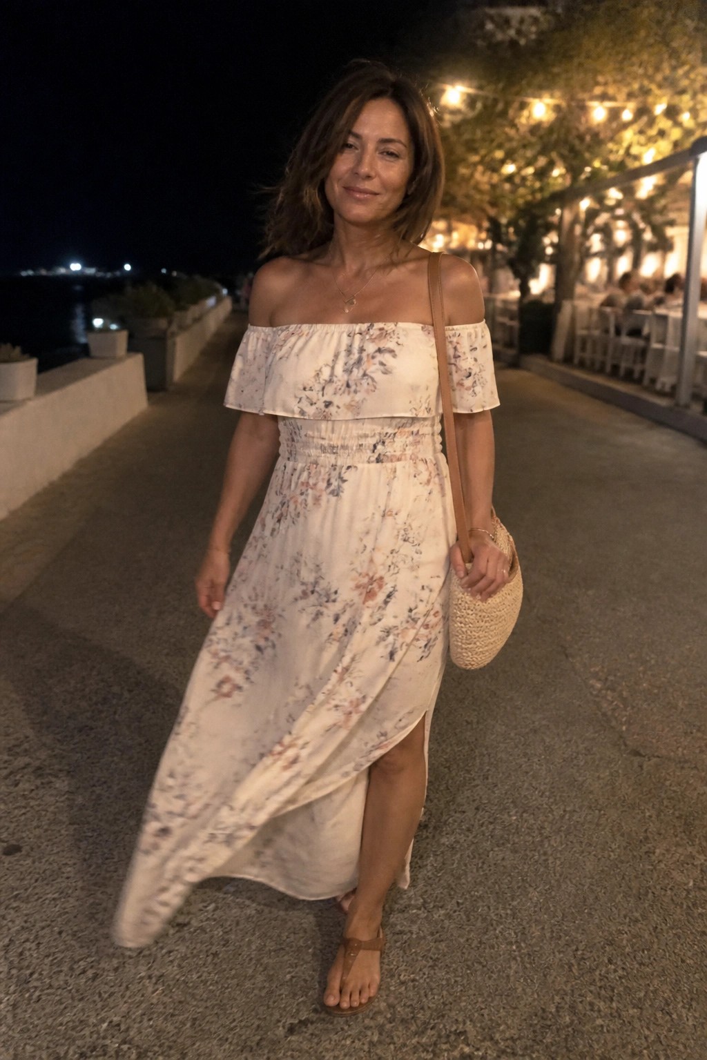 Woman wearing a white floral off-shoulder maxi dress with side slit, carrying a woven straw bag and wearing brown sandals.