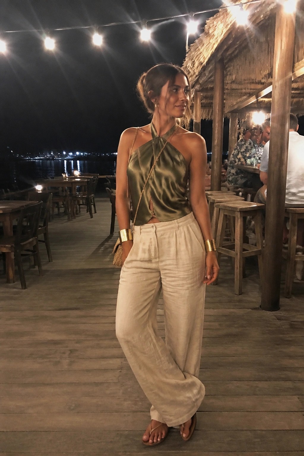 Woman in green halter top and beige wide-leg pants on lit deck at night
