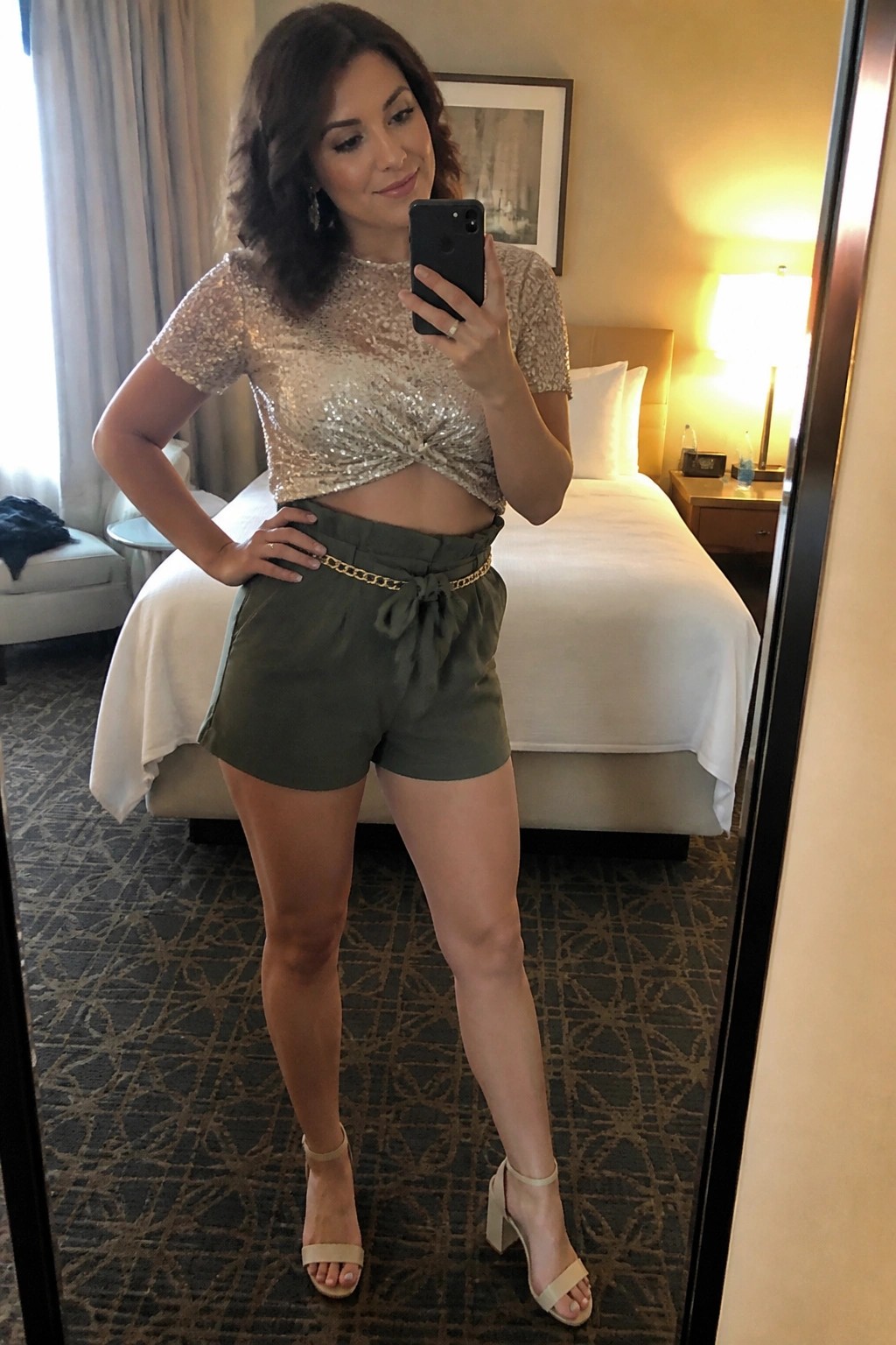 Woman in sequined crop top and olive shorts taking mirror selfie in hotel room