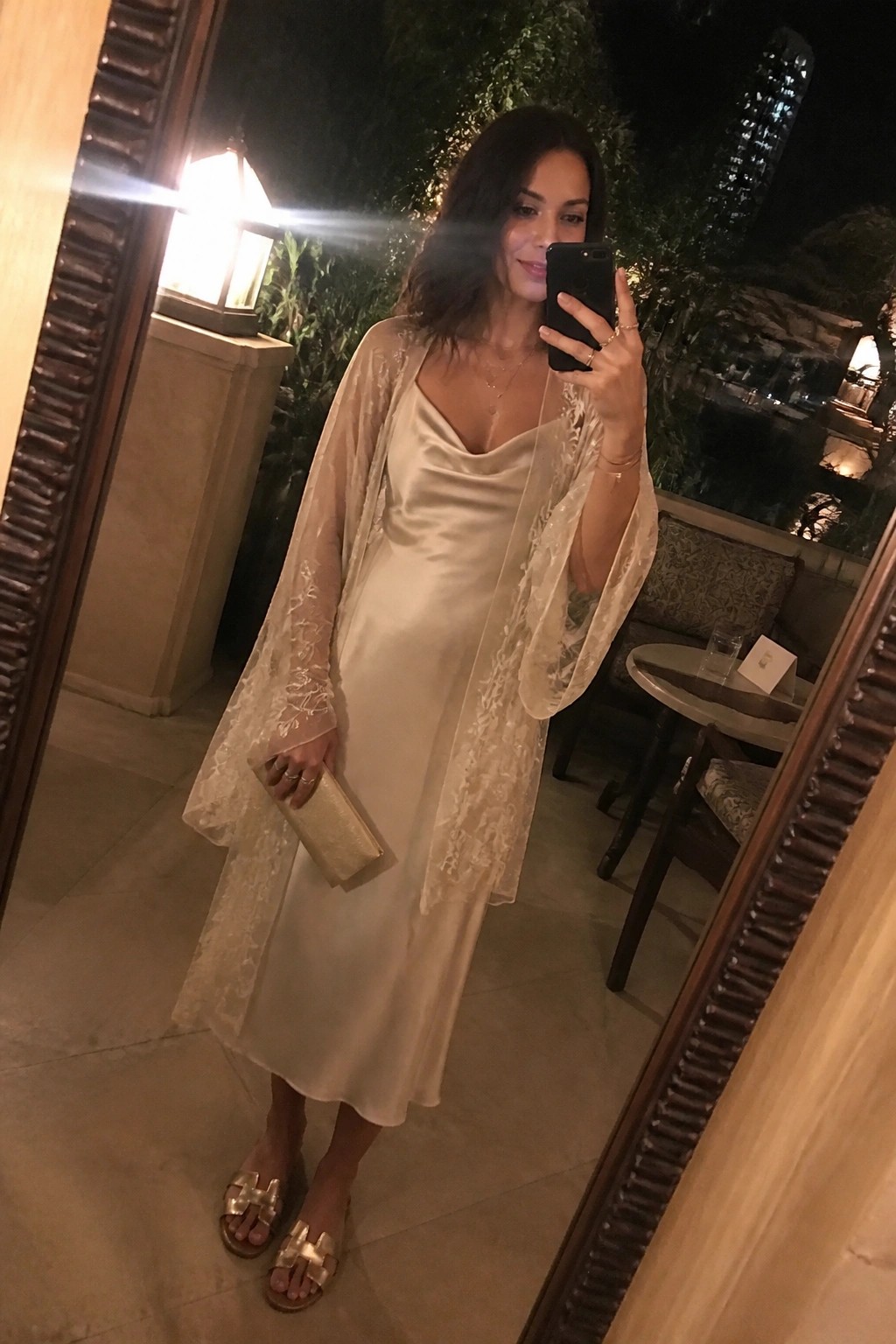 Woman wearing an ivory satin slip dress layered with a sheer embroidered lace robe, holding a gold clutch and wearing gold sandals.