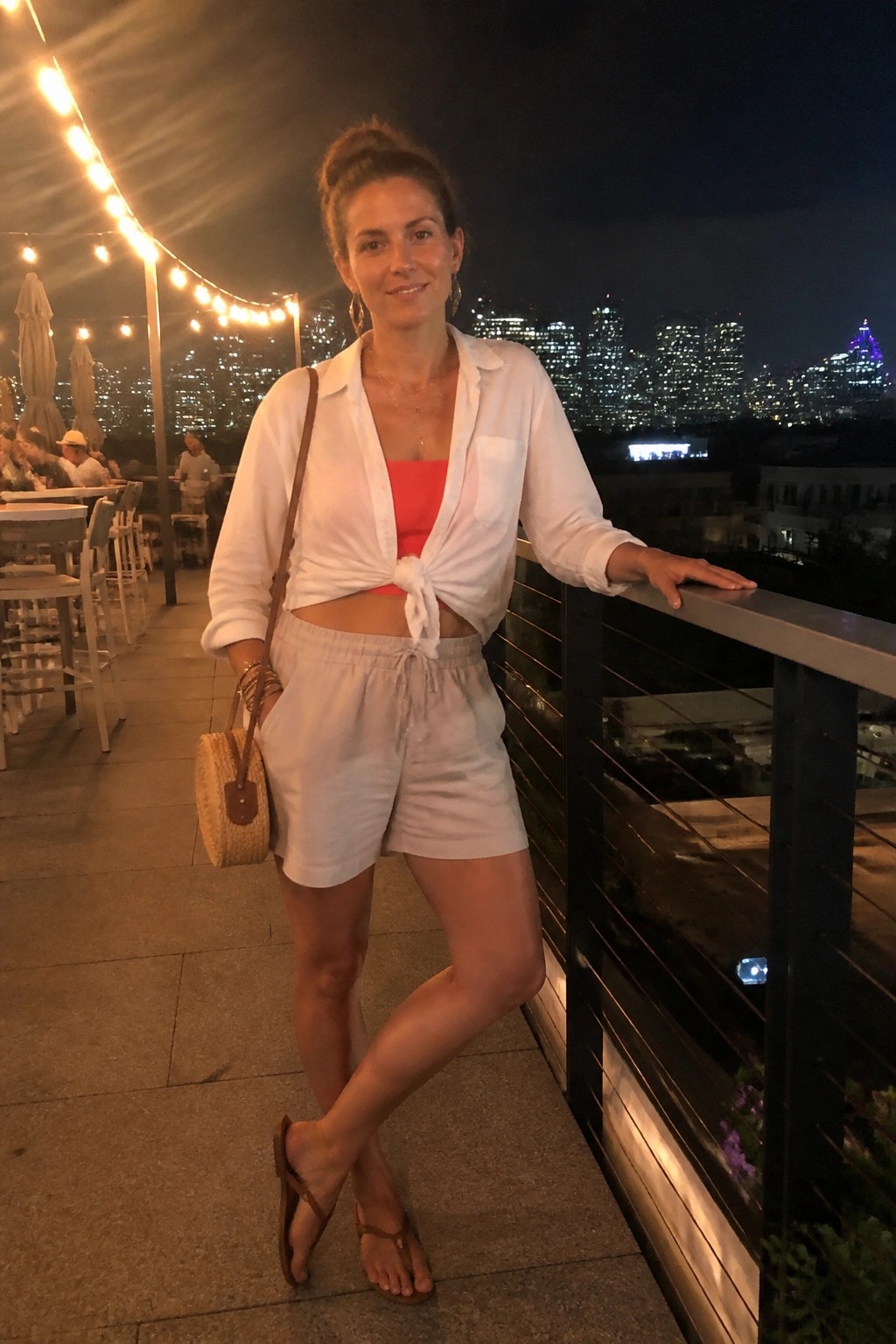Woman standing on a balcony at night wearing a white button-up shirt tied at the waist over a coral crop top, paired with light gray drawstring shorts, brown flip-flops, a round woven crossbody bag, layered necklaces, and bracelets, with a city skyline visible behind her.