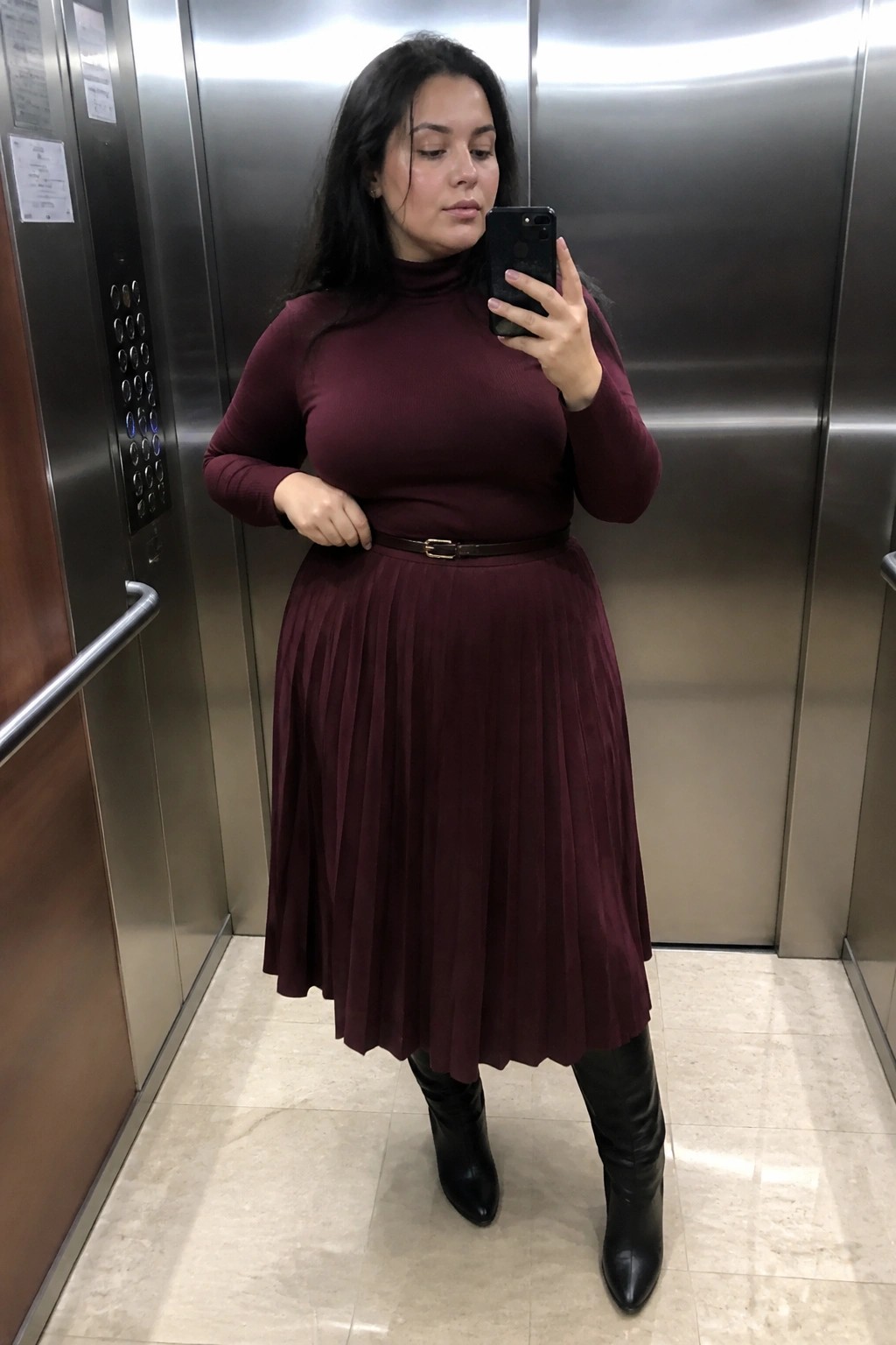 Woman in maroon turtleneck and pleated skirt taking elevator selfie