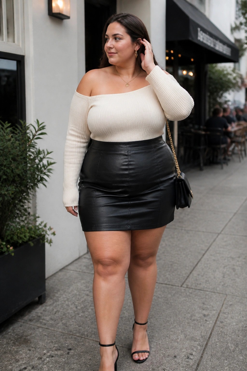 Plus size woman in cream off shoulder ribbed sweater tucked into black leather mini skirt paired with black chain strap bag and strappy heels.